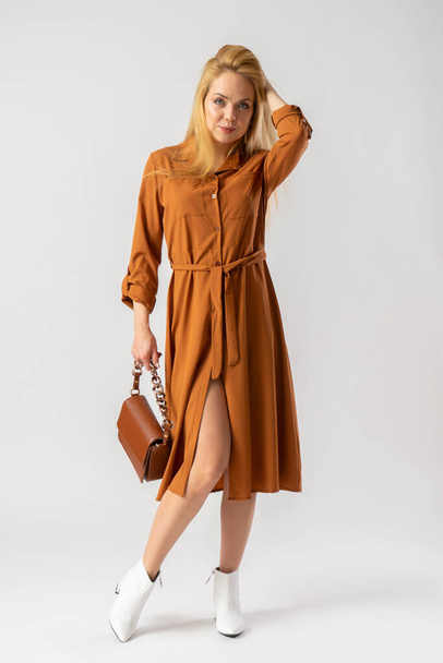A young blond woman in an elegant casual brown dress, with a small brown leather handbag. Model posing in the studio on a white background. - Foto, afbeelding