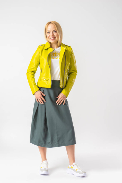 Молодая блондинка в стильной юбке, ярко-зеленой кожаной куртке и белой футболке. Фотомодель позирует в студии на белом фоне. - Фото, изображение