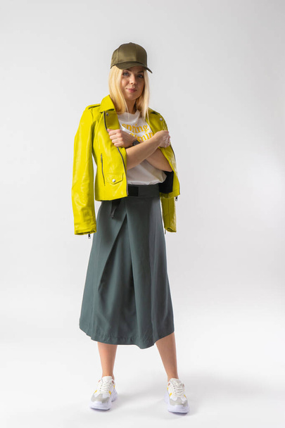 A young blonde woman in a stylish youth casual skirt, a white T-shirt and a baseball cap threw a light green leather jacket over her shoulders. Model posing in the studio on a white background. - Zdjęcie, obraz