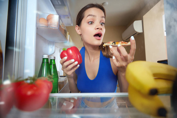 Young woman opening fridge, looking inside and emotionally choosing what to eat. Pizza and vegetables. View from inside of fridge. Concept of food, diet, emotions, domestic lifestyle, taste - Photo, Image