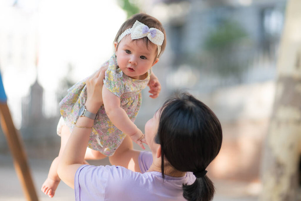 outdoors lifestyle portrait of Asian Chinese woman holding beautiful and adorable baby girl smiling cheerful on playground wearing cute ribbon in mother daughter bonding concept - Foto, imagen