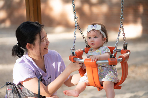 outdoors lifestyle portrait of Asian woman playing with beautiful and adorable baby girl smiling cheerful on playground playing on swing at city park wearing a cute ribbon in childhood concept - Foto, imagen
