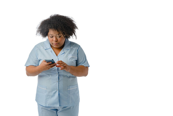 joven mujer médica afro-latina de etnia venezolana con uniforme azul, de pie revisando los mensajes en su teléfono, fondo blanco con espacio para copiar. - Foto, Imagen