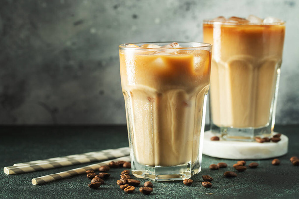 Ice coffee in a tall glass with cream poured over, ice cubes and beans on a dark concrete table. Cold summer drink with tubes on a black background with copy space. - Photo, Image