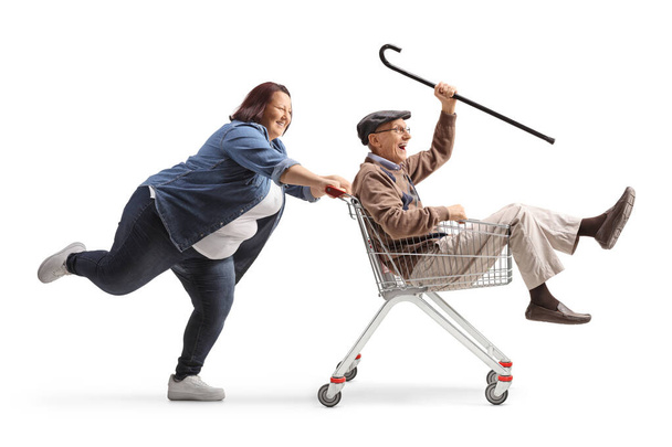 Full length profile shot of a woman pushing a senior man with a cane inside a shopping cart isolated on white background - Fotografie, Obrázek