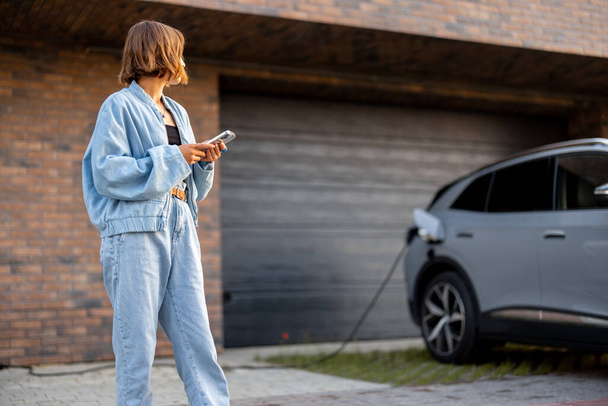 Jonge vrouw die een smartphone gebruikt terwijl ze haar elektrische auto oplaadt in de buurt van de garage van haar huis. Begrip technologie en EV-auto 's - Foto, afbeelding