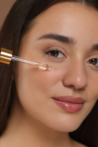 Beautiful woman applying essential oil onto face on brown background, closeup - Φωτογραφία, εικόνα