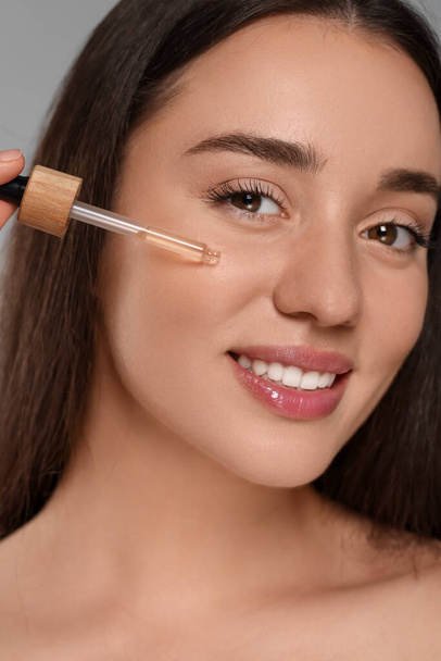 Beautiful young woman applying essential oil onto face on grey background, closeup - Φωτογραφία, εικόνα