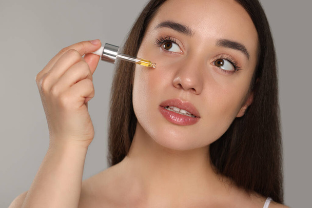 Beautiful young woman applying essential oil onto face on grey background - Фото, зображення