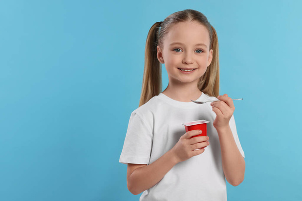Cute little girl with tasty yogurt on light blue background, space for text - Fotó, kép