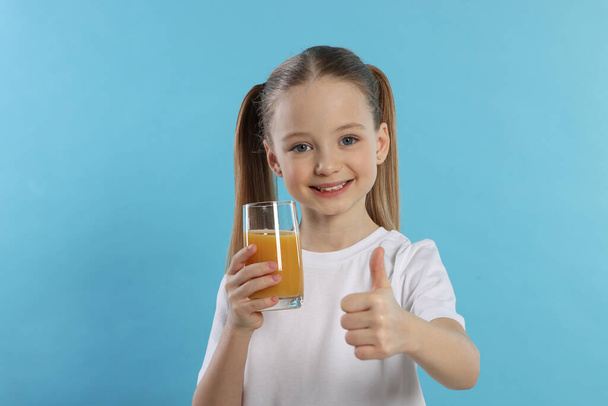 Cute little girl with fresh juice showing thumbs up on light blue background - Foto, imagen