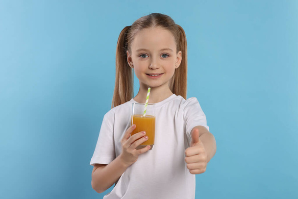Cute little girl with fresh juice showing thumbs up on light blue background - Φωτογραφία, εικόνα