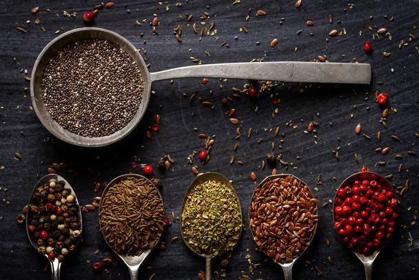 Old spoons with various seeds, herbs and spices on black background. Flat lay. Top view. Food concept. Dark mood food photography. - 写真・画像