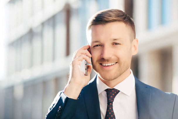 Outdoor shot of prosperous successful unshaven businessman wears formal clothes, has pleasant talk with business partner via cell phone, discuss future meeting, poses against blurred background - Photo, Image