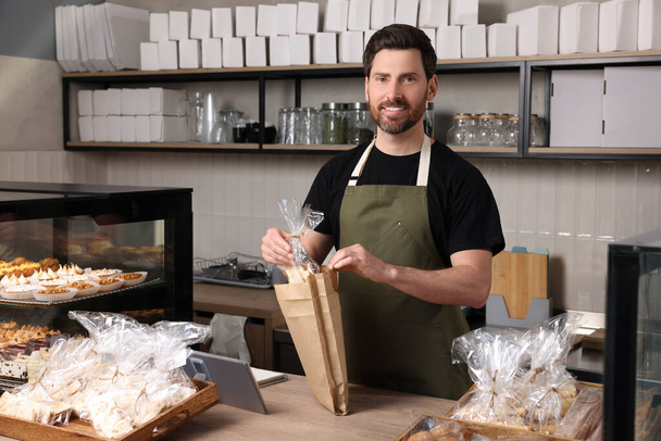 Happy seller putting pastry into paper bag at cashier desk in bakery shop - Φωτογραφία, εικόνα