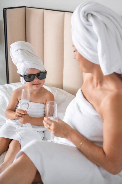 Happy family mother and little daughter in dressing gowns and towels laugh and drinking water with glasses during spa day at home. High quality photo - Foto, imagen