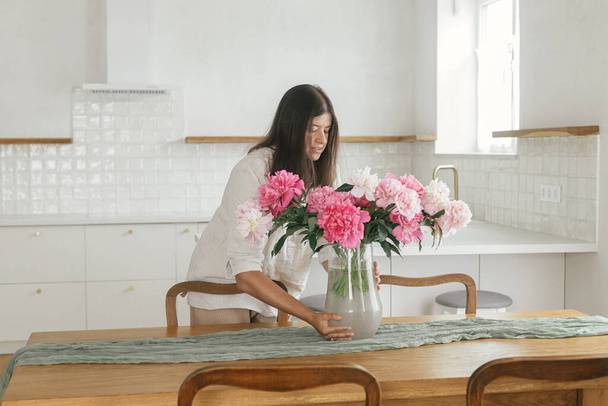 Woman arranging beautiful peonies in vase in new modern home. Young female decorating house with flowers on background of minimal white kitchen, moving in scandinavian house - Photo, Image