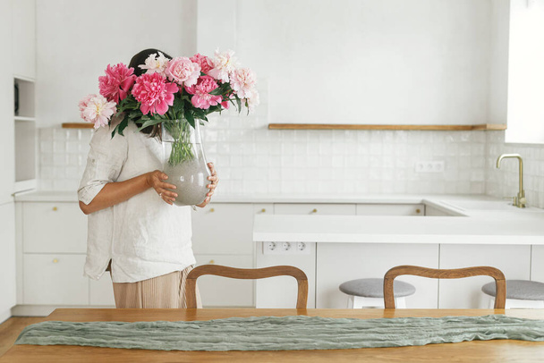 Woman arranging beautiful peonies in vase in new modern home. Young female decorating house with flowers on background of minimal white kitchen, moving in scandinavian house - Photo, image