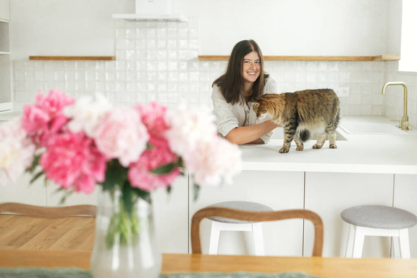 Mujer feliz acariciando gato lindo en el fondo de la cocina blanca mínima con ramo de peonías en el nuevo hogar moderno. Ama de casa relajante después de limpiar la cocina y jugar con la mascota - Foto, imagen