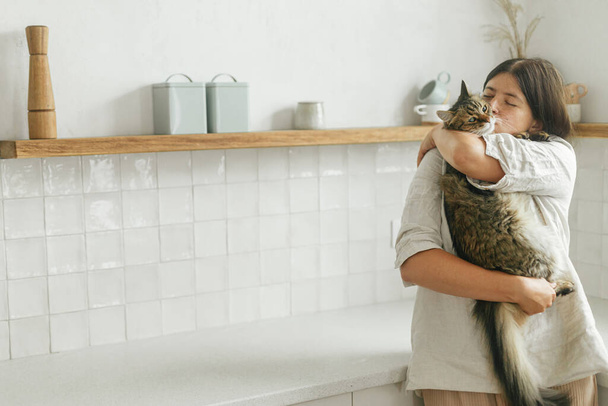 Stylish woman hugging her cute tabby cat in new minimal white kitchen. Happy female caressing and kissing adorable pet in new modern home after moving in - Fotografie, Obrázek