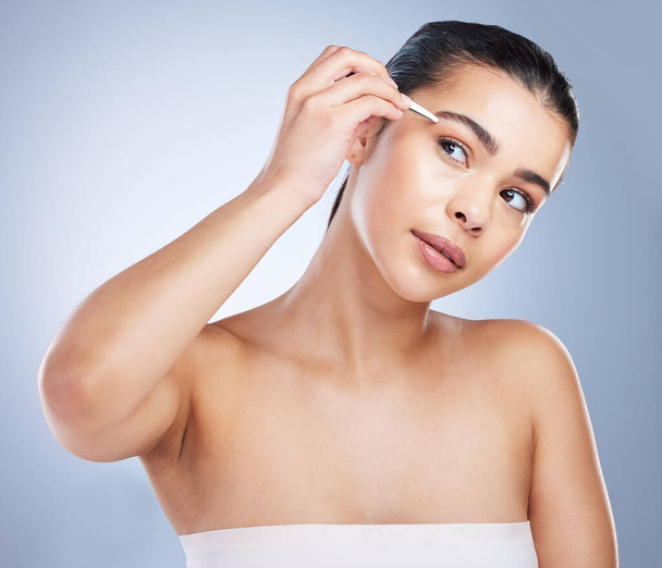 Young woman tweezing her eyebrows in a studio for grooming or hair removal face routine. Skincare, beauty and female model doing a facial epilation treatment with tweezers isolated by gray background. - Photo, image