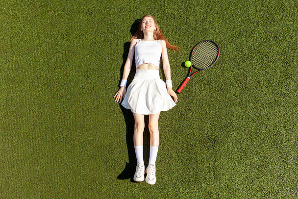 jovem jogador de tênis menina em uniforme esportivo branco com raquete de tênis mentiras e repousa sobre a quadra verde, vista superior, espaço de cópia - Foto, Imagem