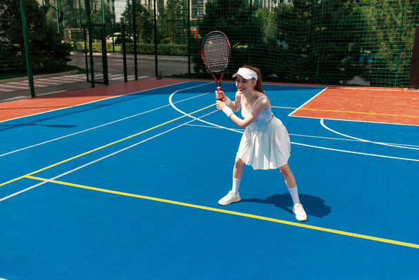 юная теннисистка в белой форме держит ракетку на теннисном корте, спортсменка играет в теннис на открытом воздухе - Фото, изображение