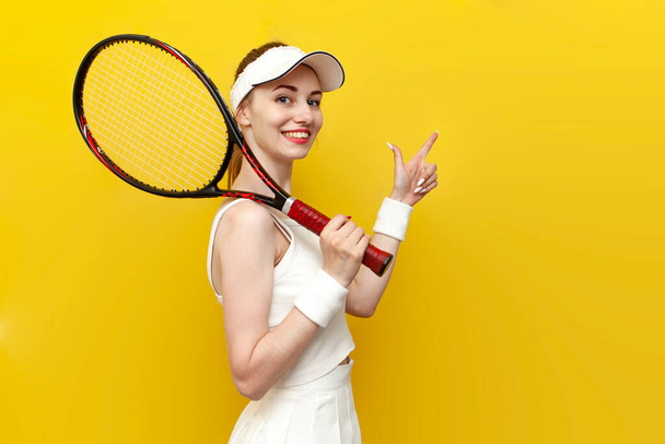 jugador de tenis chica en ropa deportiva sostiene raqueta de tenis y puntos a un lado en el fondo aislado amarillo, entrenador de tenis femenino en uniforme blanco anuncia espacio de copia - Foto, imagen