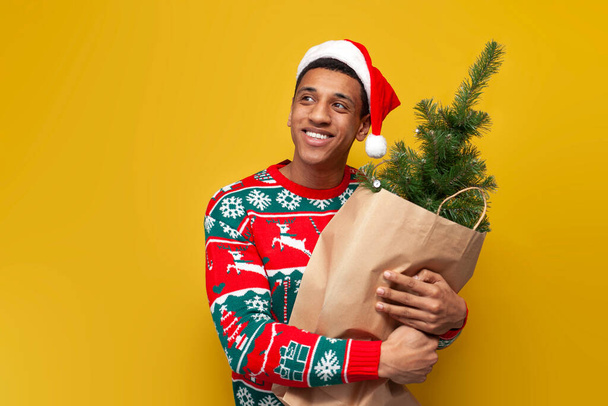 african american guy in christmas clothes and santa hat holds christmas tree in bag on yellow isolated background, man in sweater buys decorations for the new year - Фото, зображення