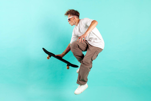 若いクレイジー男はスケートボードに乗って青い隔離された背景にジャンプし、サングラスのヒップスターは、空気中のスケートボードと飛ぶと極端なトリックを行います - 写真・画像
