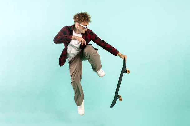 young crazy guy rides skateboard and jumps on blue isolated background, hipster in sunglasses flies with skateboard in the air and does extreme trick - Foto, imagen