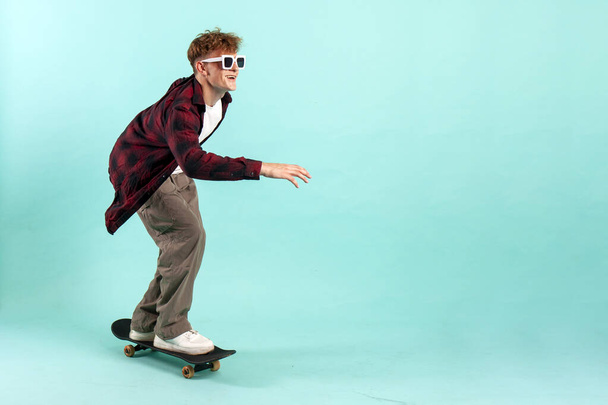 young cheerful guy rides skateboard on blue isolated background, hipster skater in sunglasses, copy space - Photo, Image