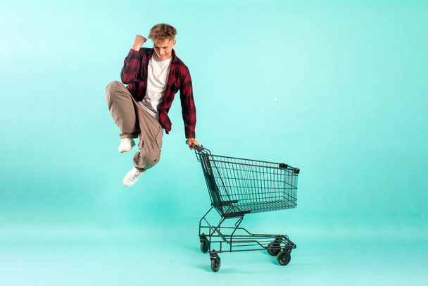 young crazy guy shopper jumps and rejoices with shopping cart from supermarket on blue isolated background, male customer quickly runs in the air for shopping - Photo, Image