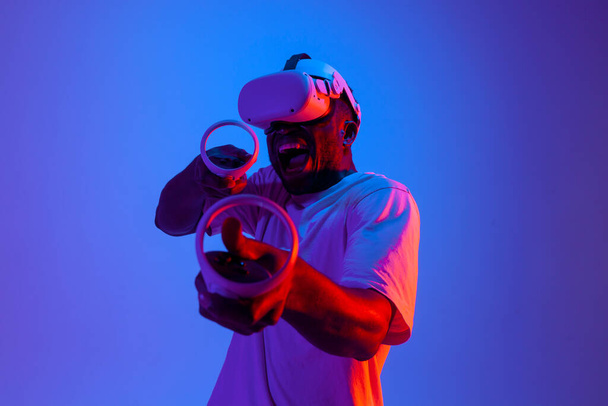 shocked african american male gamer in virtual reality glasses shoots and aims with joysticks in neon lighting, guy plays vr shooter with gadget and holds control panel and screams - Фото, зображення