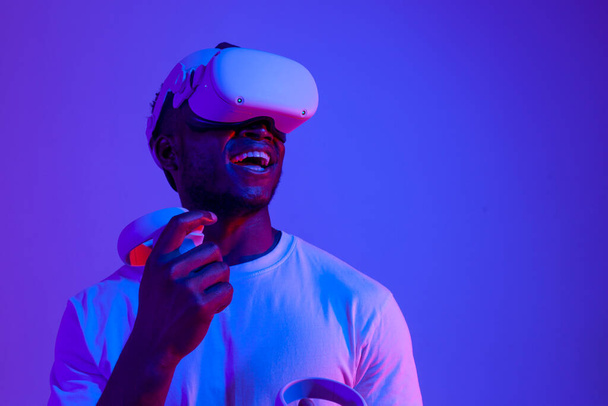 Sanal gerçeklik gözlüklü Afro-Amerikan erkek oyuncu, neon ışıkta başka yere bakıyor, VR cihazıyla oynuyor, kontrol panelini tutuyor ve fotokopi alanına bakıyor. - Fotoğraf, Görsel