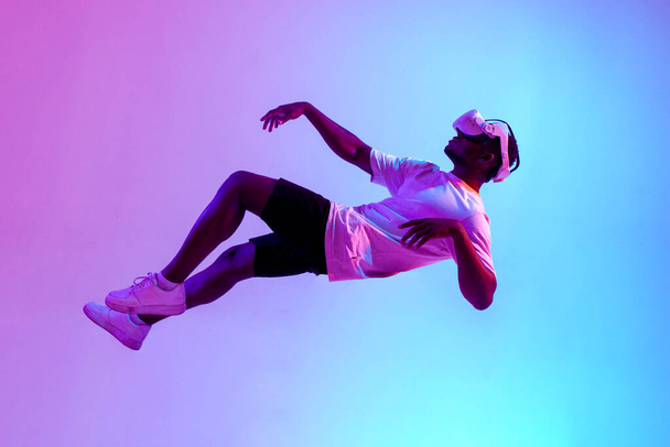 african american male gamer in virtual reality glasses flies in the air and falls in neon lighting, guy plays with vr gadget and levitates, concept of imagination and fantasy - Valokuva, kuva