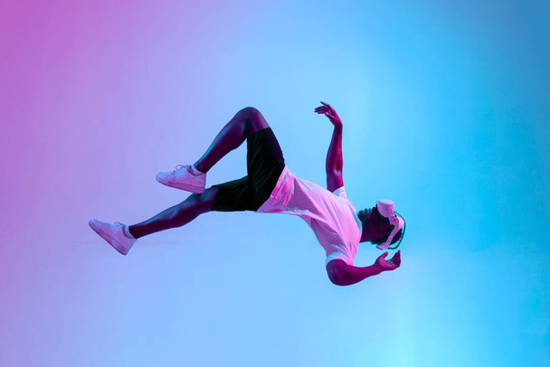 Africano americano jugador masculino en gafas de realidad virtual vuela en el aire y cae en la iluminación de neón, chico juega con vr gadget y levita, concepto de imaginación y fantasía - Foto, imagen