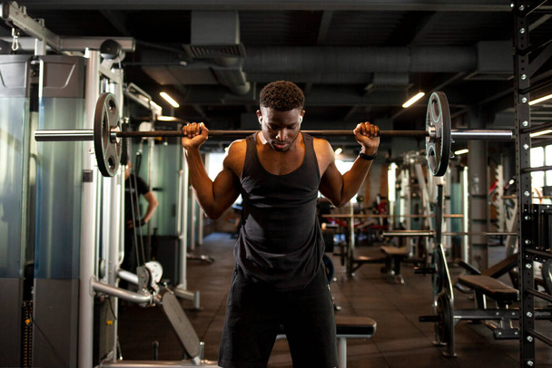 若いアフリカ系アメリカ人男性がダークジムでバーベルを持ち上げフィットネスルームでスポーツマンのトレーニングを受け - 写真・画像