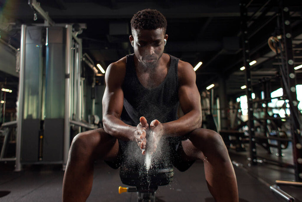 deportista afroamericano hombre entrena en gimnasio oscuro y se frota las manos con magnesia, joven chico hace aplauso con sus manos en la sala de fitness, primer plano de polvo y harina con las manos de atleta - Foto, imagen