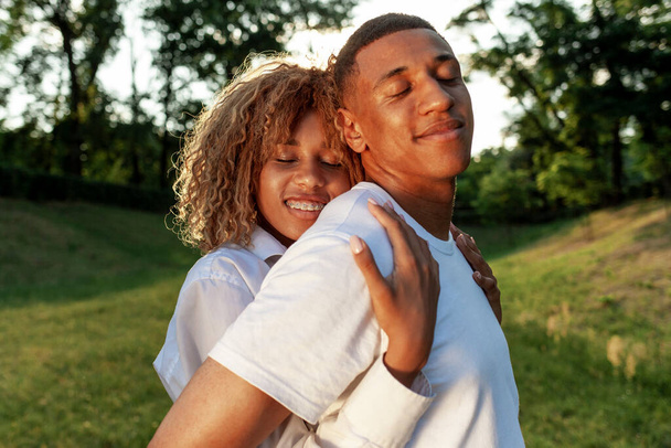 happy young african american couple standing together in the park with their eyes closed and dreaming, curly girl hugging her boyfriend and smiling, concept of love and care - Zdjęcie, obraz