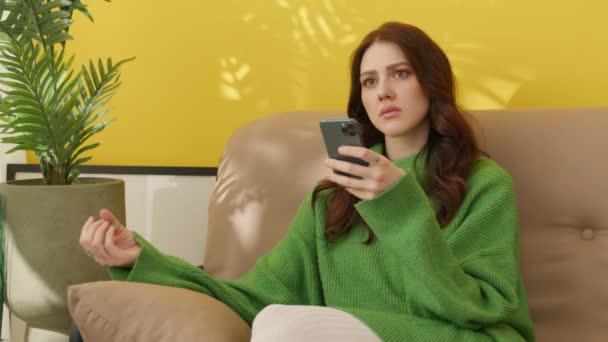 Ελκυστική νεαρή μελαχρινή με μακριά μαλλιά, light make up, πράσινο πουλόβερ, με υπερήφανη εμφάνιση πληκτρολογώντας στο smartphone κάθεται σε μπεζ καναπέ στο άνετο δωμάτιο. Υψηλής ποιότητας 4k πλάνα - Πλάνα, βίντεο