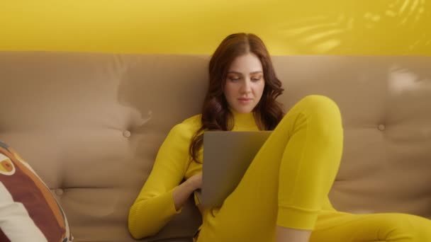 Mulher bonita nova com cachos de estilo em roupa amarela sentado no sofá com laptop, conversando. Mulher senta-se com as pernas dobradas, com a parede amarela atrás dela. Imagens 4k de alta qualidade - Filmagem, Vídeo