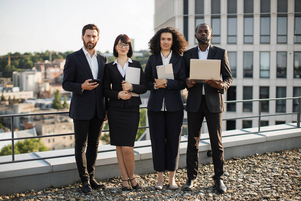 Full length portrait of four successful foreign investors dressed in business outfit posing on camera with wireless technology while standing on rooftop of office building. Concept of teamwork. - Фото, зображення