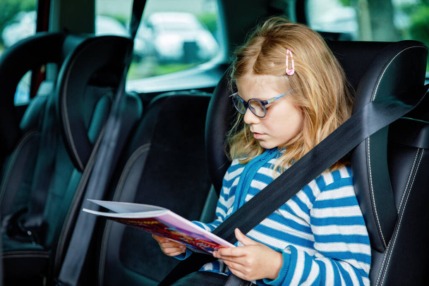 Araba koltuğunda oturan küçük anaokulu kızı. Gözlüklü mutlu çocuk kitap okuyor, trafik sıkışıklığı sırasında aile tatiline giderken gülümsüyor. - Fotoğraf, Görsel