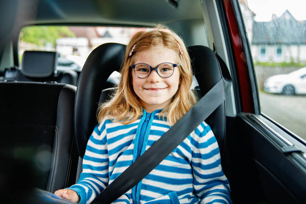 Dziewczynka z przedszkola siedzi w foteliku samochodowym. Szczęśliwe dziecko w okularach Czytanie książki, Uśmiech w drodze na rodzinne wakacje podczas korków - Zdjęcie, obraz