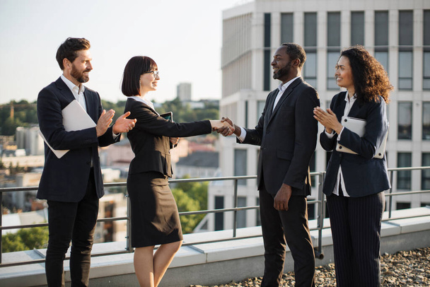 Side view of multiethnic entrepreneurs celebrating contract agreement of becoming partners. African american man and caucasian short haired lady shaking hands while their colleagues clapping on roof. - 写真・画像