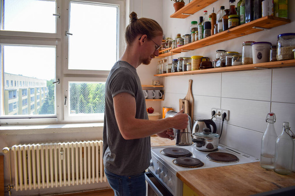 Улыбающийся молодой бизнесмен готовит кофе с итальянской кофеваркой, работая дома. Веселый мужчина-фрилансер, работающий удаленно из домашнего офиса. Налейте в чашу - Фото, изображение