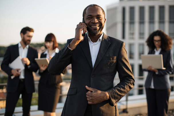 Beaming african american gentleman having conversation on smartphone during meeting with partners on terrace. Multiracial colleagues in suits using remote gadgets. Concept of business cooperation. - Valokuva, kuva