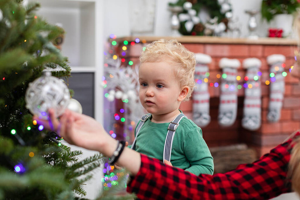 Rozmazané popředí. V popředí žena ruka ukazující cetku visící na vánočním stromečku. Vedle ní stojí malý chlapec a dívá se na cetku. V pozadí mlhavý cihlový krb - Fotografie, Obrázek