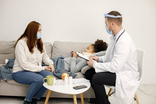 Male doctor in white medical gown asking sick girl about her symptoms. Black girl has fever and overspread blanket. Different medicines are on table near sofa. - Photo, Image